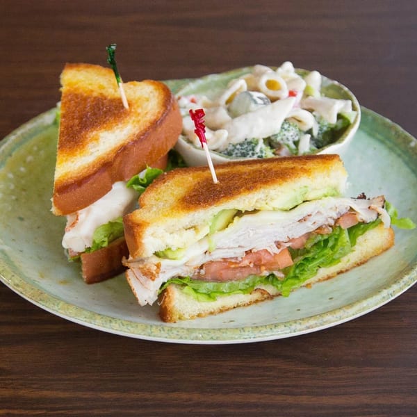 A turkey club sandwich with lettuce, tomato, and avocado, served with a side of pasta salad.