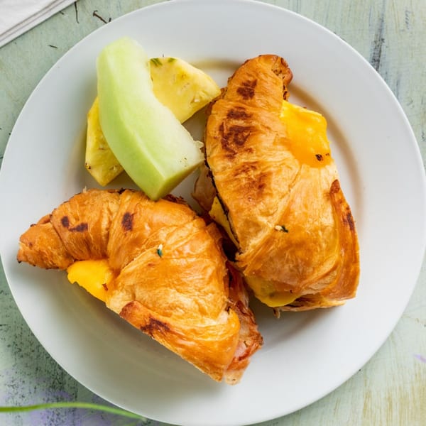 Two golden croissants filled with cheese, served with fresh pineapple and honeydew melon on a white plate.