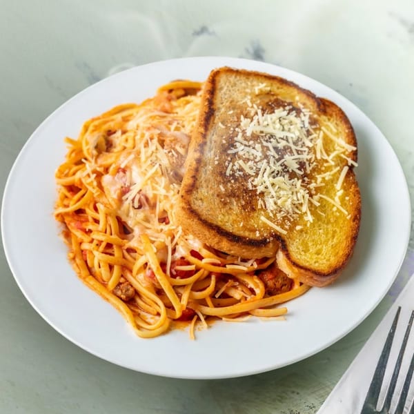 A white plate of spaghetti with meat sauce, topped with shredded cheese and a slice of garlic bread.