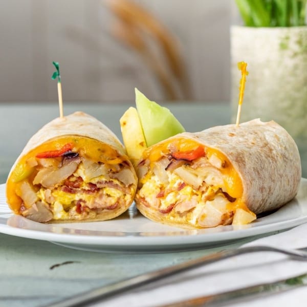 Two halves of a breakfast burrito filled with eggs, cheese, potatoes, and bacon, served with fruit.