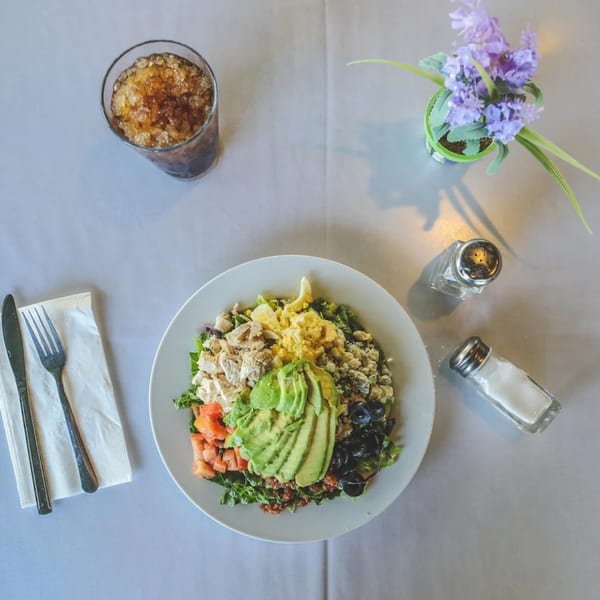 Overhead view of a Cobb salad with avocado, chicken, egg, and olives, a drink, and flowers.