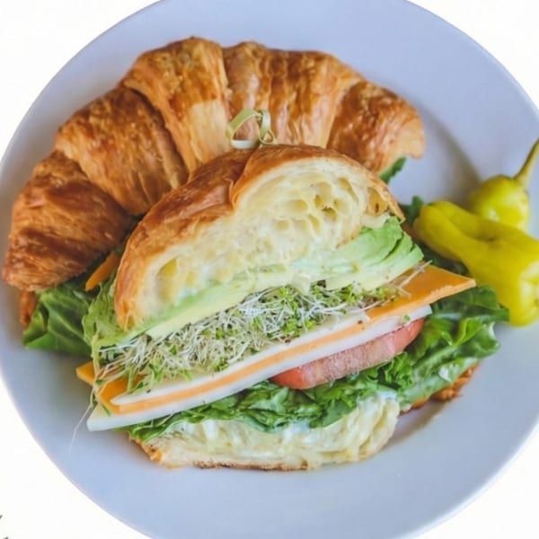 A croissant sandwich filled with lettuce, tomato, cheese, avocado, and sprouts, served with yellow peppers.