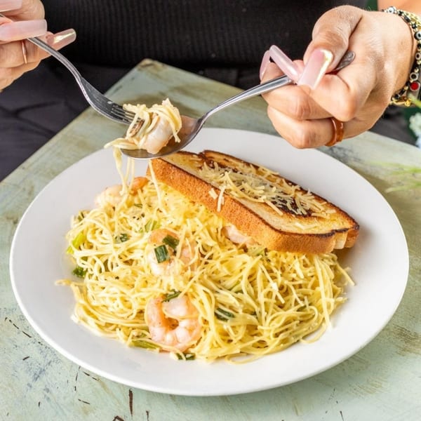 Hands twirling shrimp scampi pasta with garlic bread on a white plate at Greenhouse Cafe.